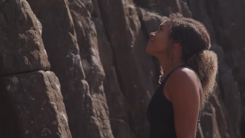 A woman stands at the base of a cliff staring towards the top