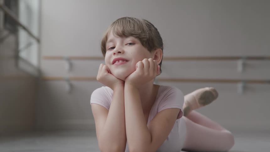 A young girl in a pink leotard and ballet slippers smiles while lying on the floor of a dance studio