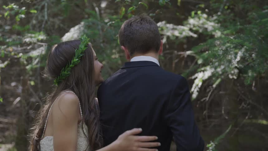 A young couple dressed in formal wear embrace and laugh in a forested area
