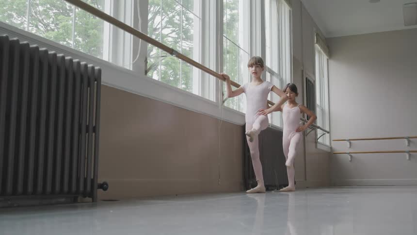 Two little ballerinas holding the handrail warm up for dancing in the ballet studio