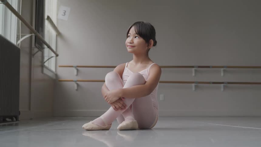 Little ballerina sits on the ground and smiles in the ballet studio