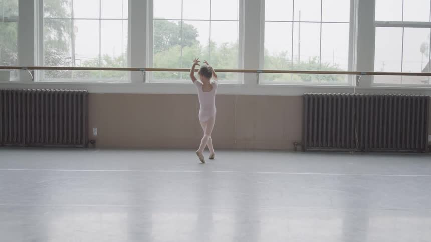 A young ballerina puts her arms above her head and twirls around in front of a ballet barre and windows in a dance studio
