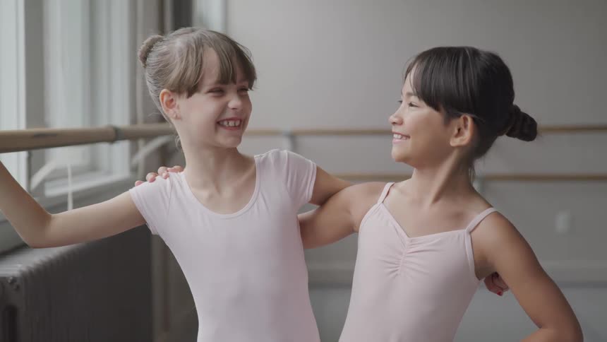 Two young ballerinas are holding onto one another and smiling while one holds a ballet barre in a dance studio