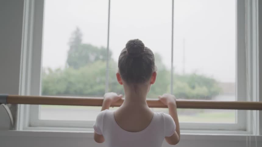 A young ballerina holds onto the barre and looks out a window