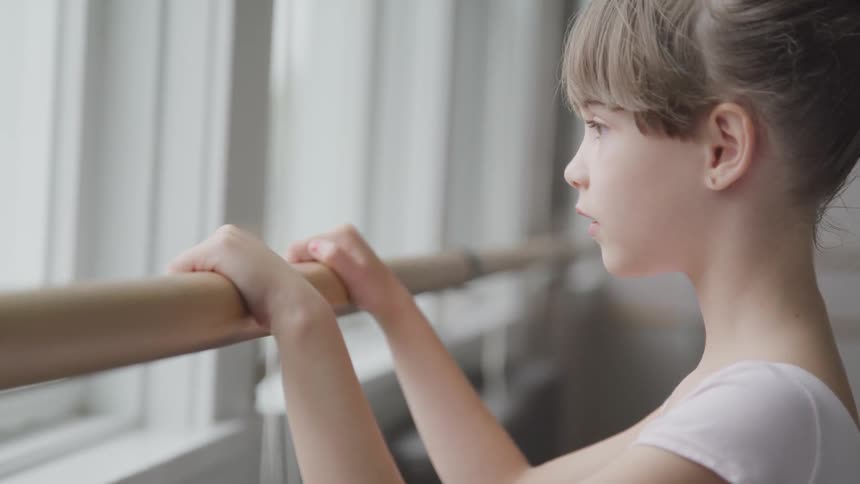A young girl stands at a ballet barre in a classroom