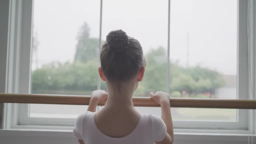 A young ballerina withher hair in a bun stands in front of a ballet barre looking out the window
