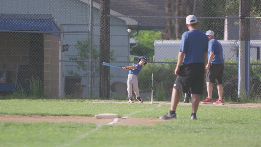 A young boy in a softball uniform swings a baseball bat and hits a baseball from home plate on a baseball diamond