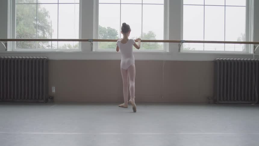 A young ballerina holds on to a dance bar by large windows