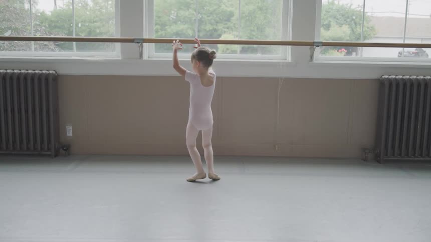 Female child ballerina in the dance studio wearing a pink leotard and ballet shoes has her arms up over her head and twirls around in a circle on her tip toes