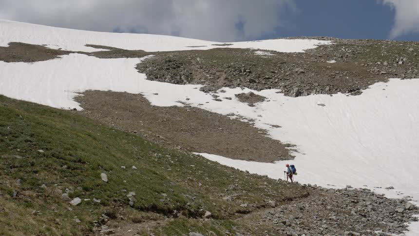 A hiker rests on their poles at the base of a slope with snow and rock behind them