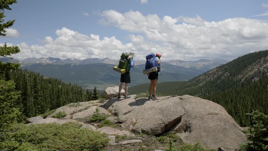 A man and woman stand on top of a cliff with their backpacks