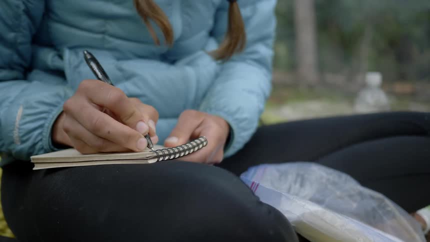 Female sitting by a campfire writing in a notebook during the day