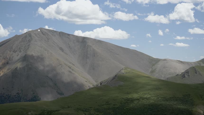 A small hill sits under a large mountain sitting under a blue sky with puffy clouds