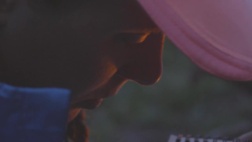 A man in a hat sits and looks at a notebook as his face is illuminated by a nearby campfire