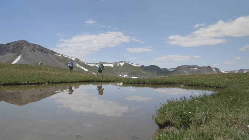 Two hikers with large backpacks stride along side a pond with a large mountain range looming in the background
