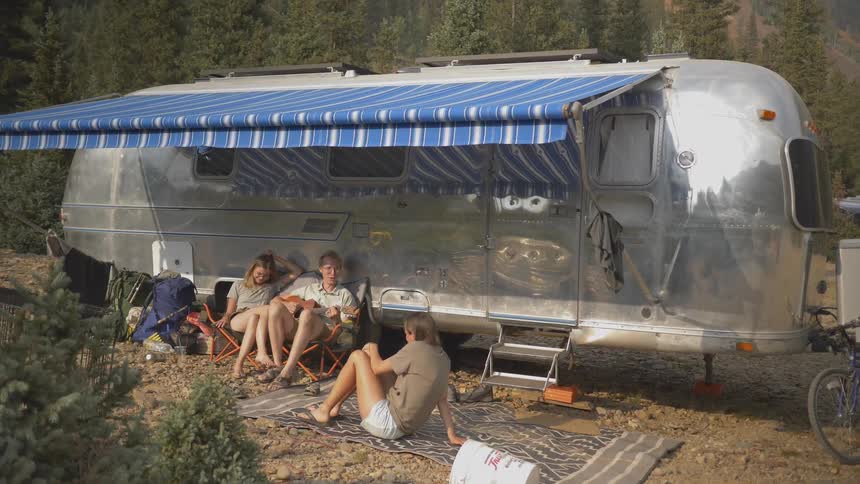 A groups of young people relax outside of a metallic vintage camper trailer parked in a forest
