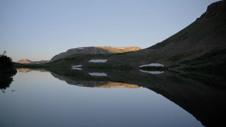 Still lake reflects slope surrounding slopes vegetation and clear blue sky