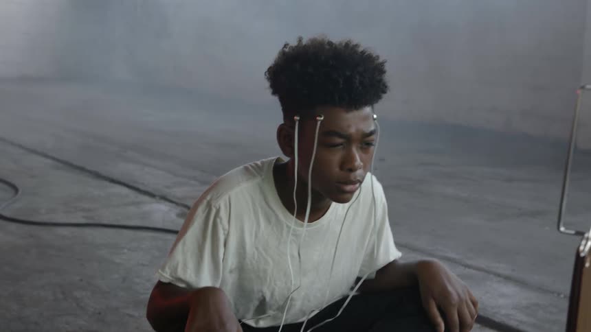 A boy sits on a cement floor with nodes connected to his head watching a small television