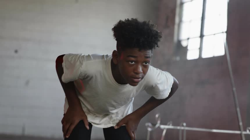 While staring straight ahead a young black male in a concrete room leans over a small television with dozens of wires hanging out the back of it