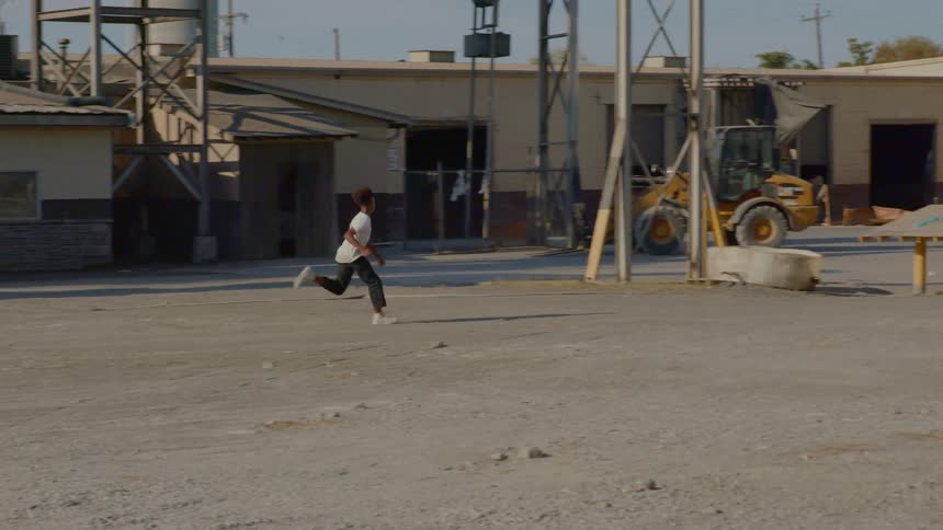 A black teen runs through an industrial complex with a dirt lot