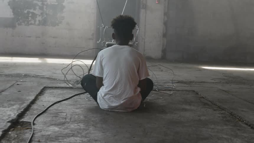 A young black teenager sits on the concrete floor of an empty room looking at a display that is surrounded with wires