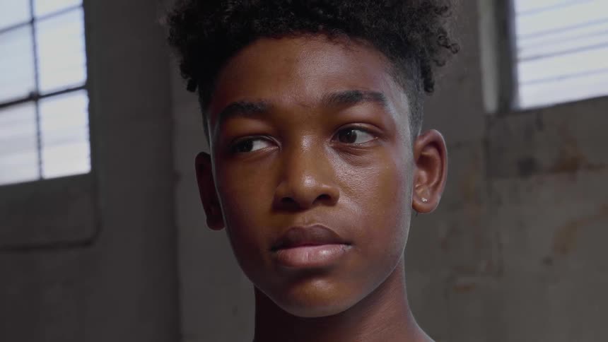 A young black teen with shifting eyes stands in front of a wall