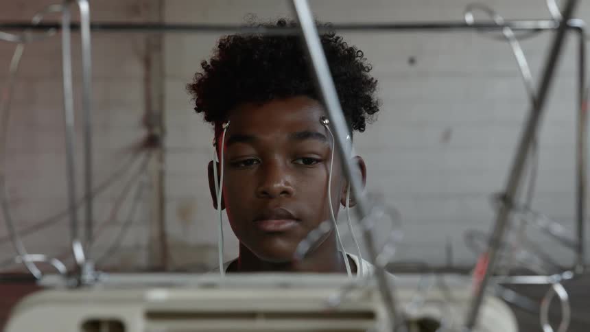 A young black teenager has wires attached to his head as his eyes dart back and forth while staring at a television