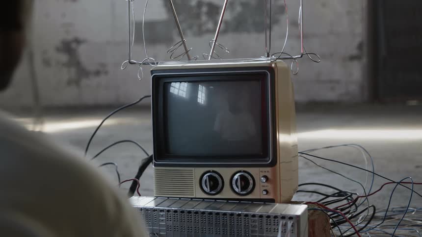 A person sits in front of an old analog tv that has dials to change the channels and antennas and wires all around it in an empty room
