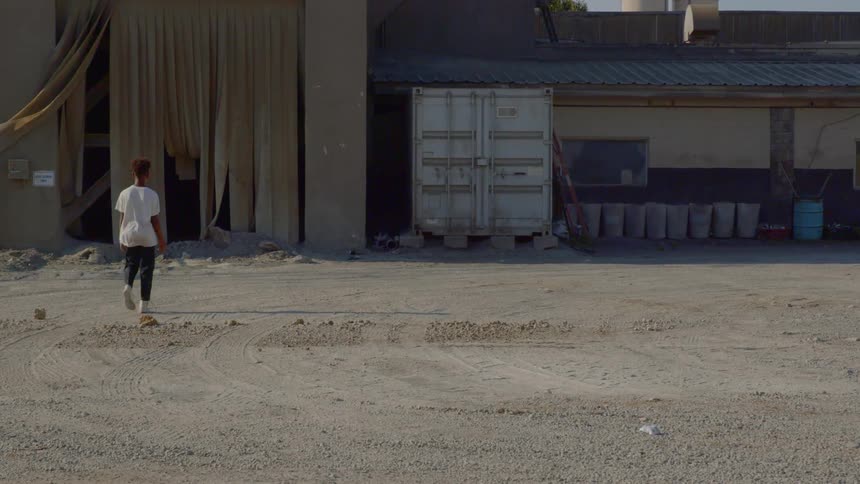 A person walks through an empty dirt parking lot towards a warehouse
