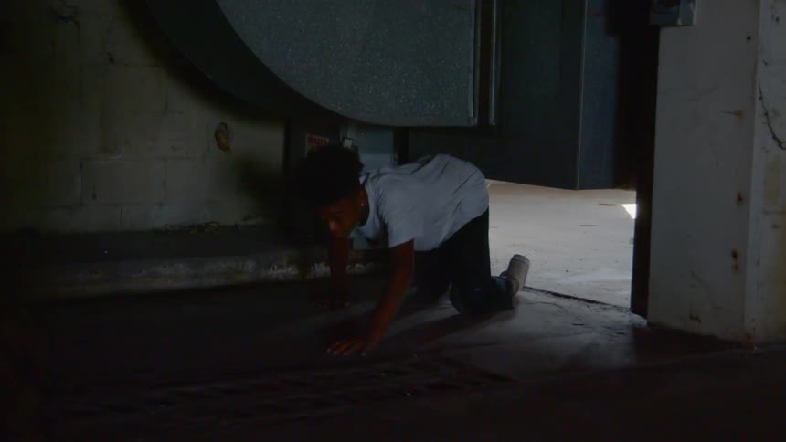 A young black male crawls underneath an air duct and then gets up to run through a room with flickering light
