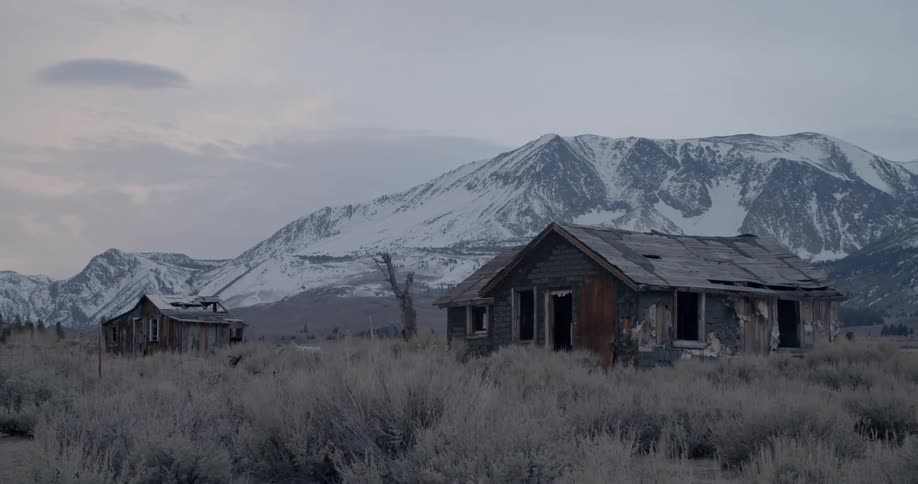 Two vacant and rundown cabins sit in a grassy field in front of snowy mountains on a cloudy day