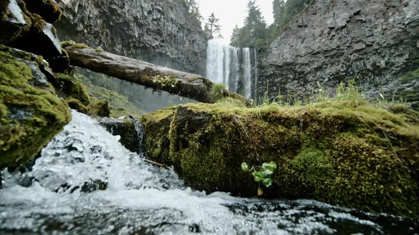 A river flows downhill between moss covered boulders while a waterfall streams down the side of a cliff in the background