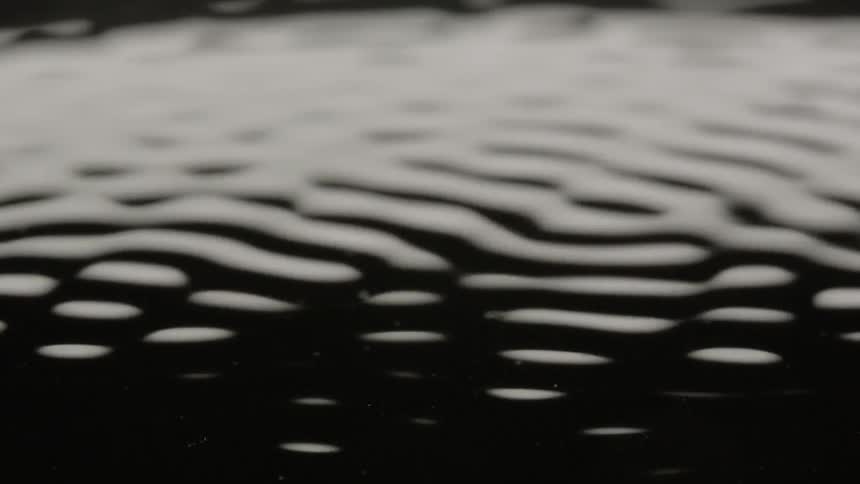 The surface of a dark black fluid is disturbed which causes numerous ripples to propagate across the surface as if being vibrated