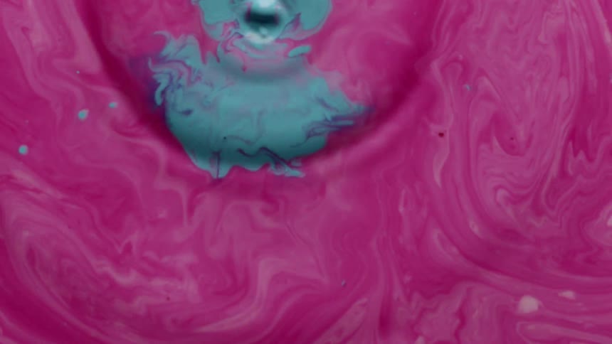 The surface of a pink liquid is disturbed by turquoise droplets that fall into it causing circular ripples