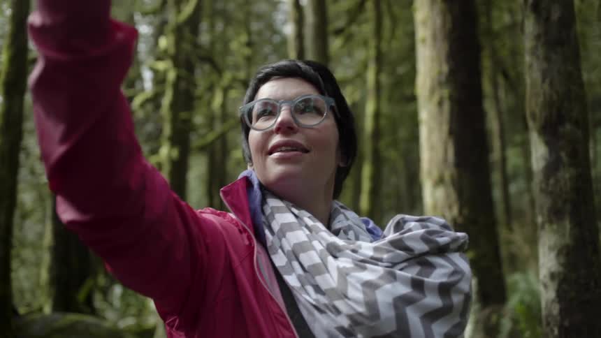 A woman stands with her arm outstretched to take a selfie in the woods
