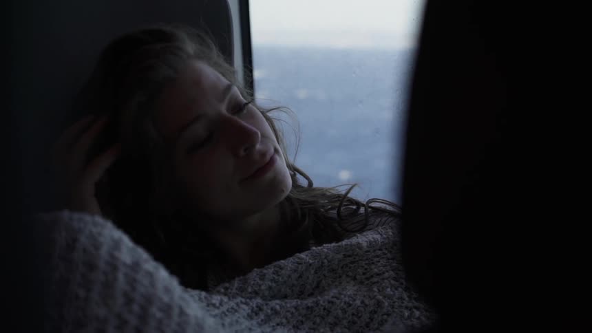 A woman with long curly hair is curled up under a blanket next to a window through which the ocean can be seen being passed by quickly