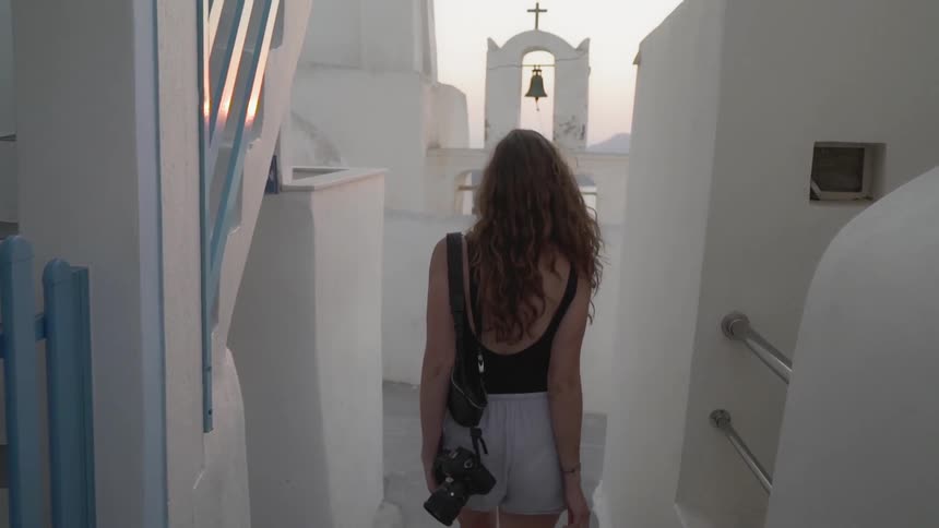 A woman carrying a camera walks down steps past white walls toward a bell tower at sunset