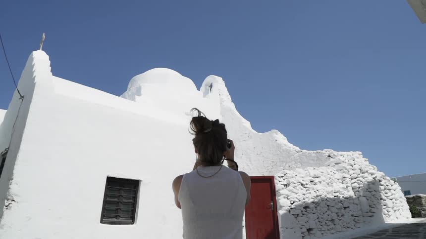 A woman stands amidst several pure white brick buildings and take a photo of one with her camera