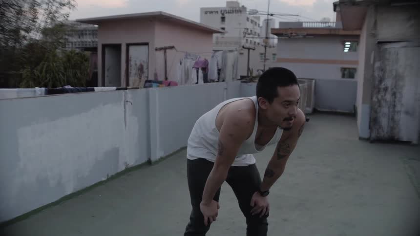A man stands on a rooftop and rests his arms against his legs before jumping around