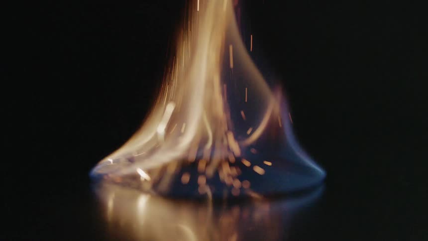A spark ignites a circular deposit of flammable liquid atop a dark reflective surface