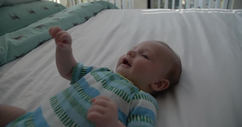 Two hands tease a happy baby that is laying on a bed