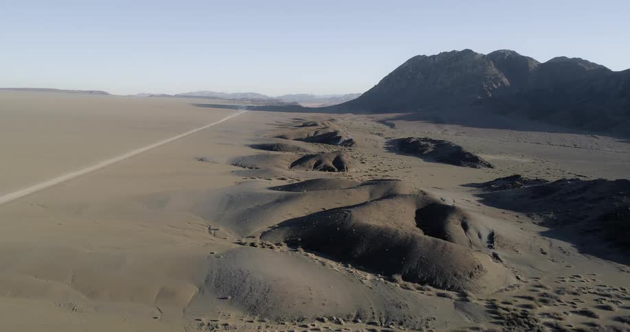 A long dirt road lies between a smooth portion of desert from some large sand dunes and mountains