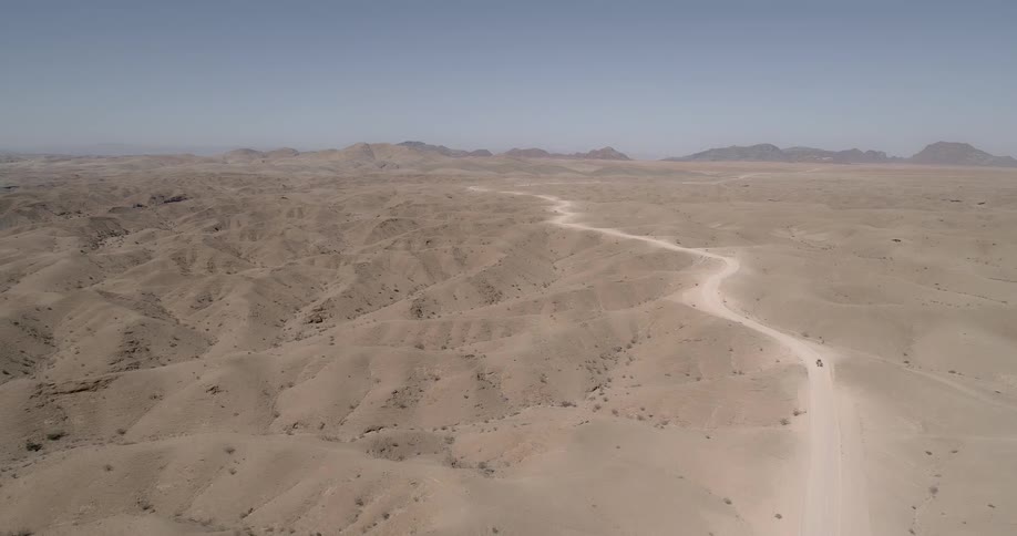 A vehicle makes its way along a long dirt road that zigzags across an expansive desert landscape