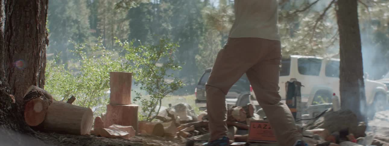 A man in tan pants swings an ax high above his head before bringing it down to chop a piece of wood at a campsite