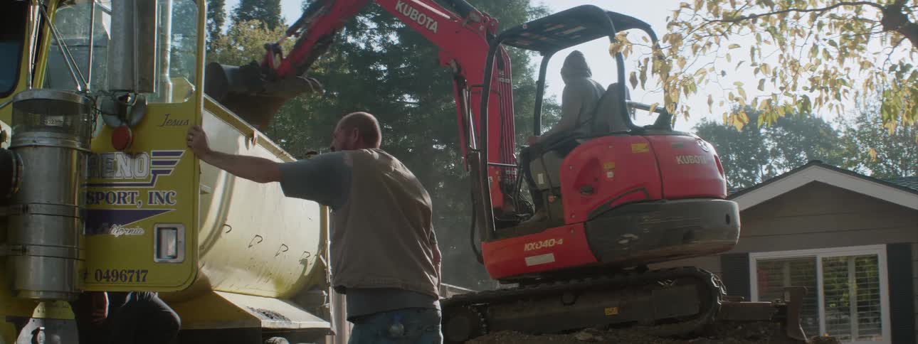 A backhoe operator turns arm away from a dump truck and back towards a yard
