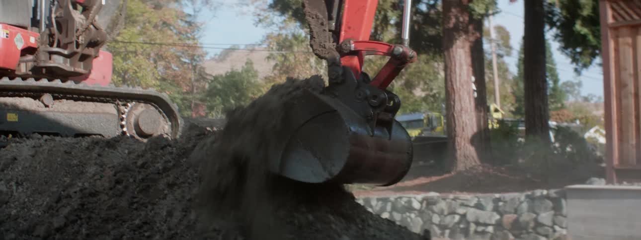An earth mover shovels moves dirt while a worker sprays it with a hose