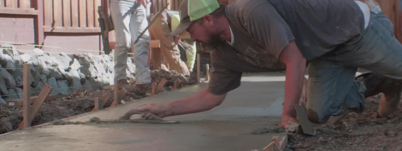 A worker leans over a freshly poured concrete walkway and smooths it using a tool