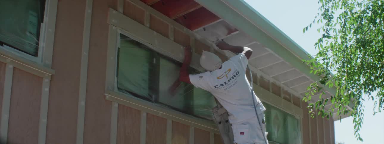 A man wearing a protective mask on his face is on a ladder using a paint sprayer to paint the overhangof a home