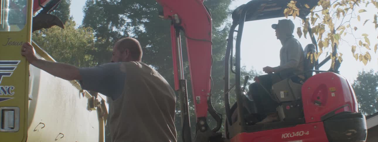 A man operating a back hoe loads dirt into a nearby truck