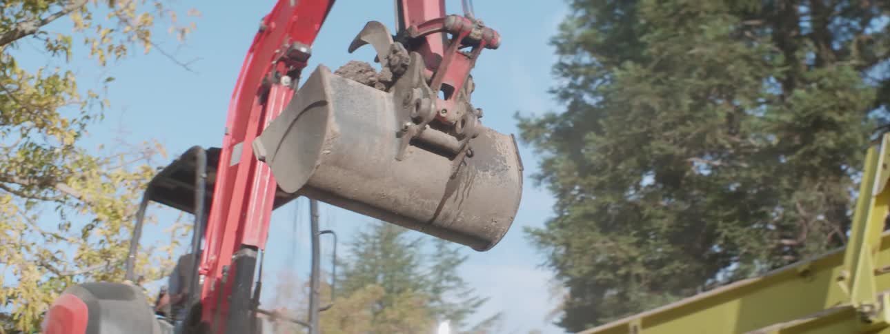 A construction worker hoses down a dirt pile while a piece of equipment scoops it up and dumps it
